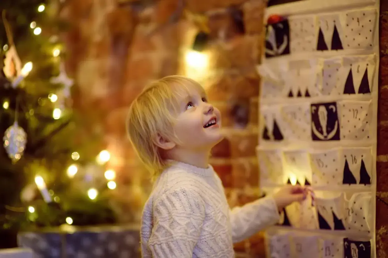 Kleiner Junge freut sich über Adventskalender mit Grundschule Ideen. Weihnachtsbaum und Lichter im Hintergrund.