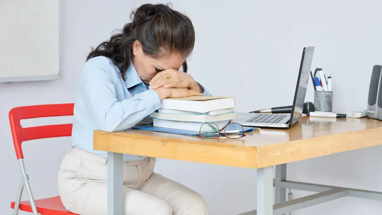 Erschöpfte Frau am Schreibtisch, fragt sich: "Soll ich Lehrer werden?" Bücher, Laptop und Brille liegen bereit.