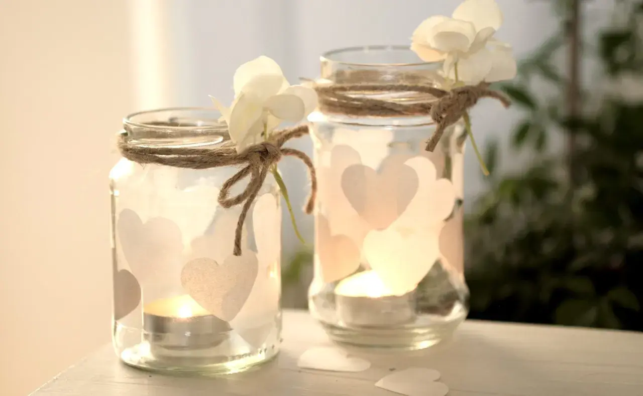 Zwei selbstgemachte Windlichter aus Gläsern, verziert mit Herzen und Blumen, strahlen warmes Kerzenlicht aus. Perfekt zum Windlichter basteln!