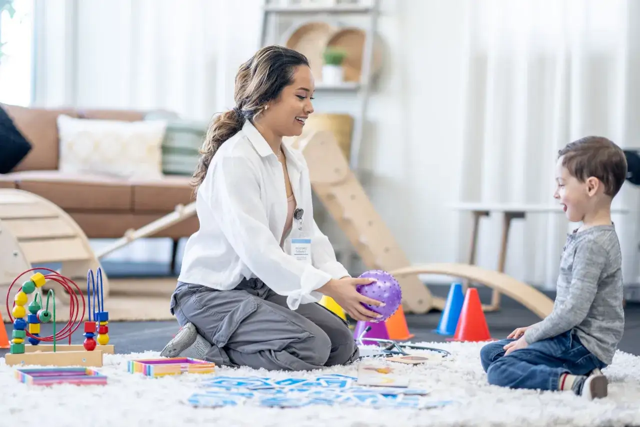 Ergotherapie bei ADHS: Eine Therapeutin spielt mit einem Kind auf einem Teppich, um Konzentration und Feinmotorik zu f&ouml;rdern. Bunte Spielsachen liegen herum.
