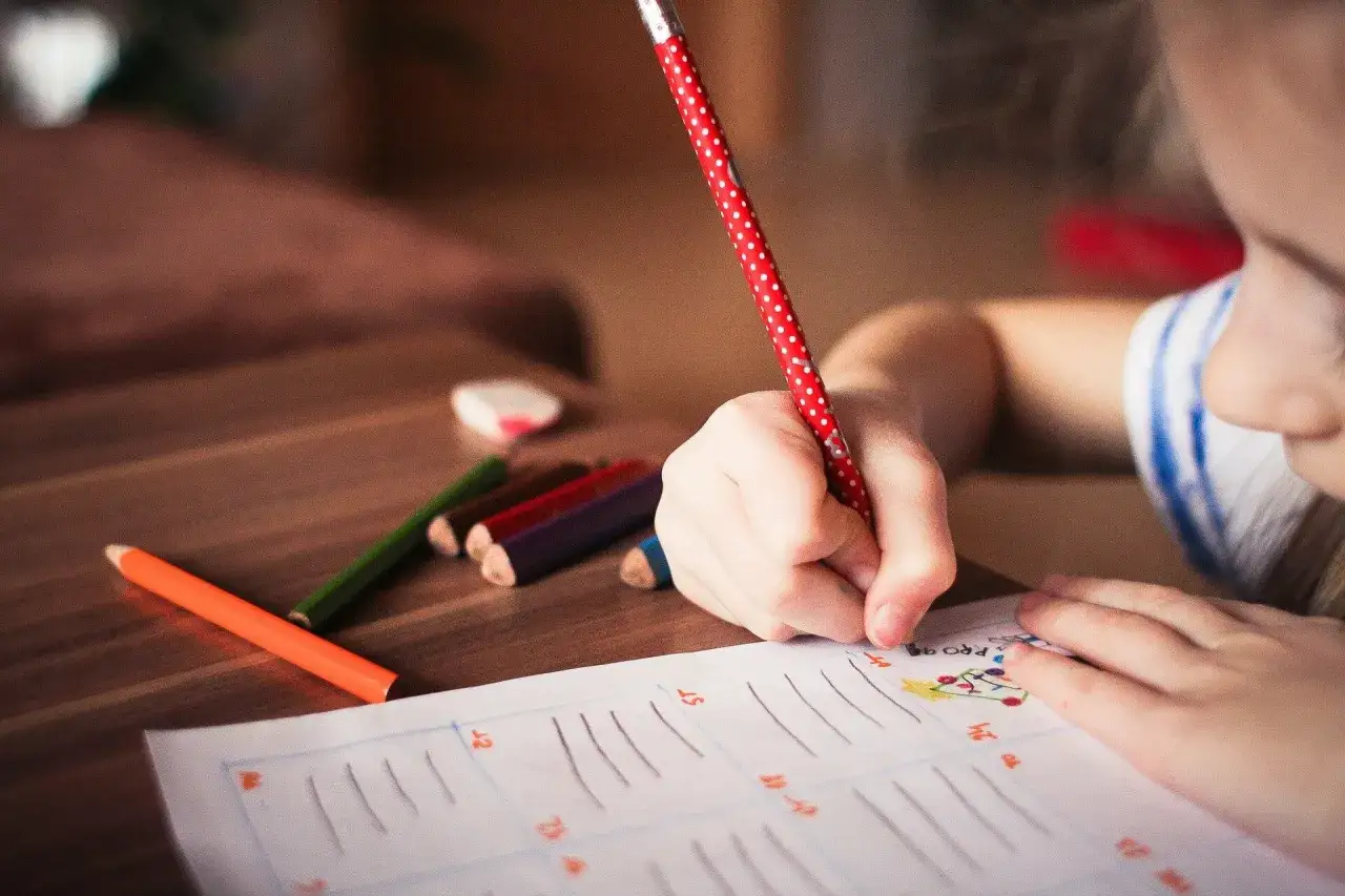 Kind &uuml;bt Graphomotorik: Mit einem gepunkteten Stift malt es auf einem Arbeitsblatt. Buntstifte liegen bereit.