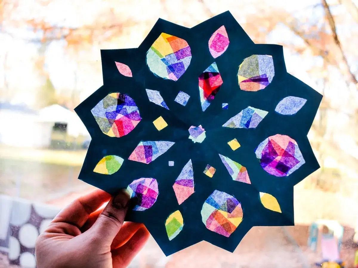 Bunte Schneeflocken basteln Kindergarten: Ein Stern aus Papier mit bunten Seidenpapierst&uuml;cken, gehalten von einer Hand vor einem Fenster.