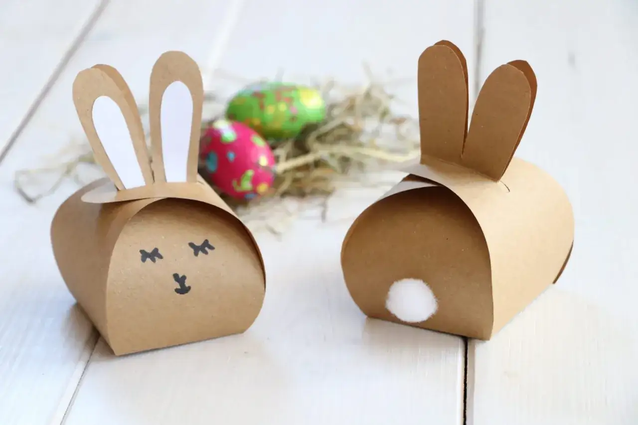 Zwei süße Papier-Osterhasen zum Basteln, mit Ohren und Schwänzchen. Im Hintergrund bunte Ostereier im Nest.