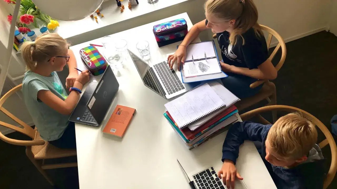 Drei Kinder lernen zu Hause, was homeschooling bedeutet. Sie sitzen an einem Tisch mit Laptops und Büchern.