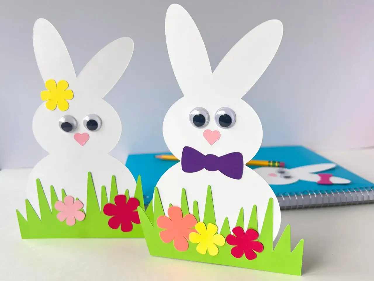 Zwei s&uuml;&szlig;e Osterhasen aus Papier, die Kinder leicht basteln k&ouml;nnen, stehen im gr&uuml;nen Gras mit bunten Blumen.