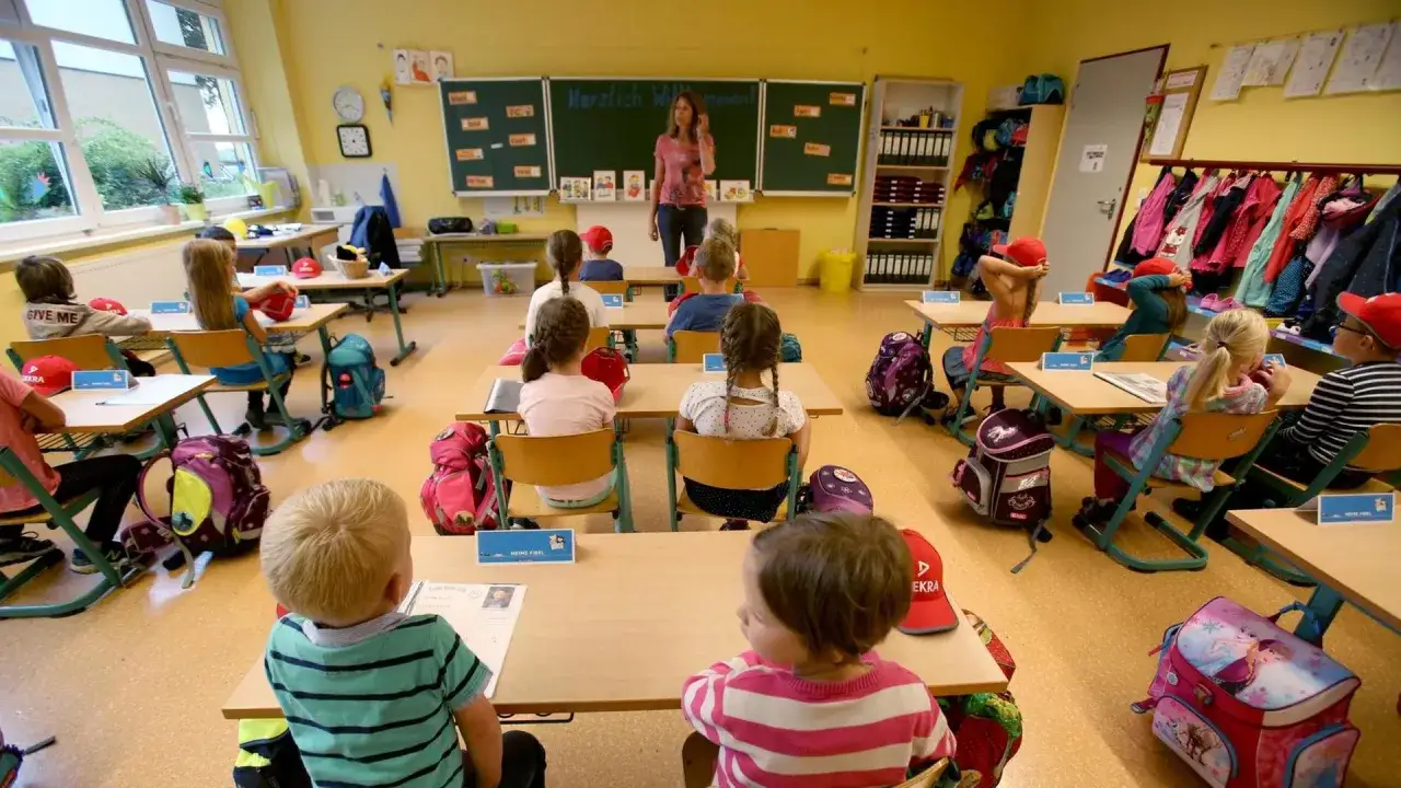 Kleine Kinder sitzen in einem Klassenzimmer. Die Lehrerin steht vorn. Soll ich Lehrer werden?