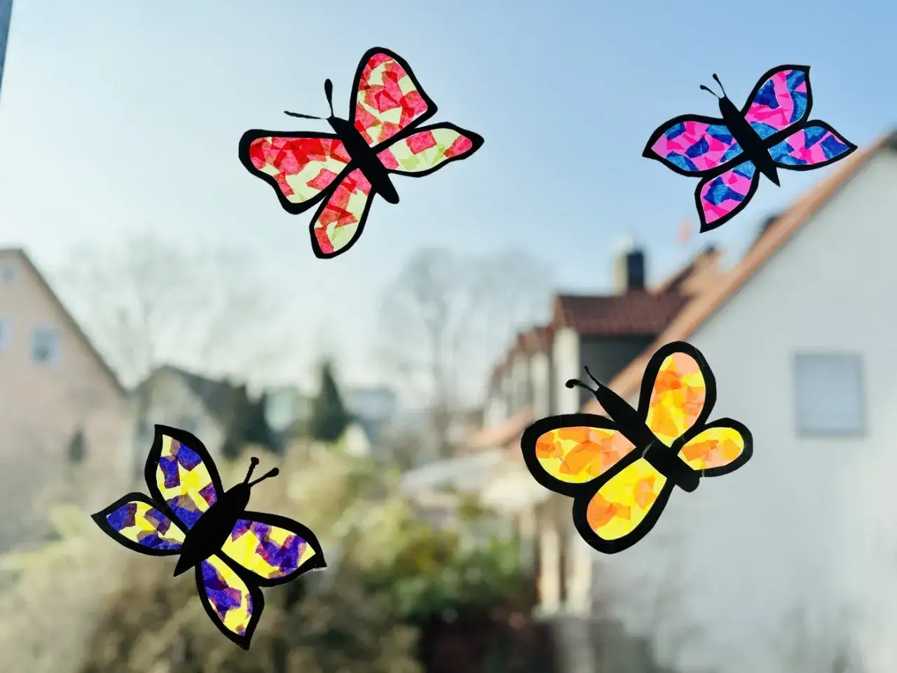 Vier bunte Schmetterlinge, selbstgemacht zum Thema Fensterbilder basteln, flattern vor einem Haus im Sonnenschein.