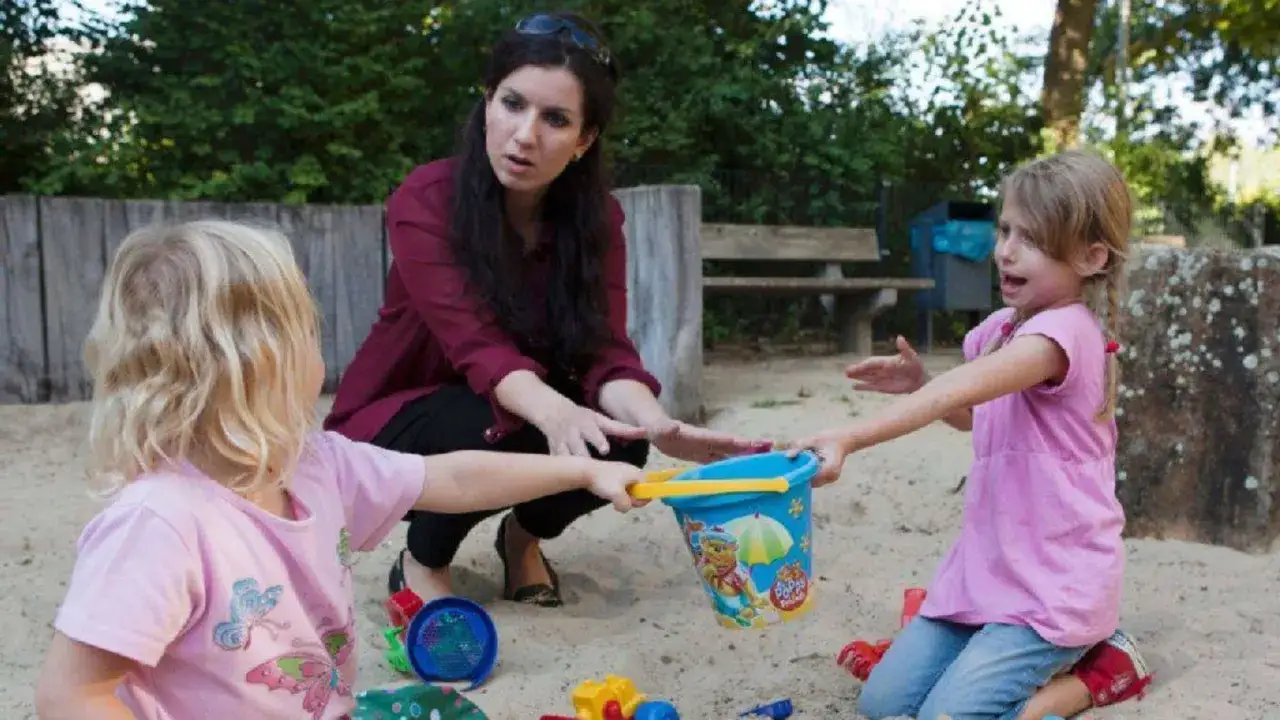 Erzieherin interveniert bei aggressivem Verhalten bei Kindern im Kindergarten. Ein M&auml;dchen versucht, ein Sandeimer wegzunehmen, w&auml;hrend die andere versucht, ihn zu verteidigen.