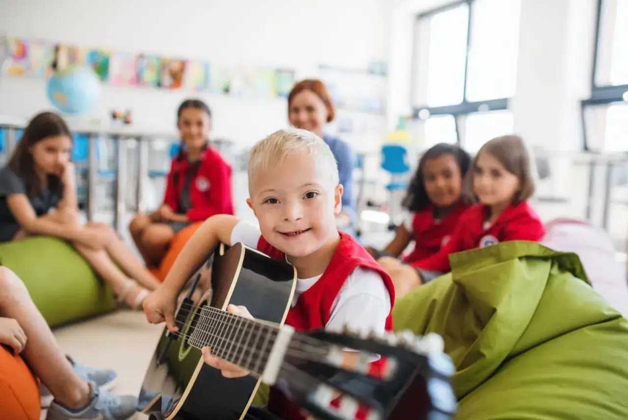 Ein Junge mit Down-Syndrom spielt Gitarre in einer Klasse. Die Lehrerin und andere Kinder schauen zu. Die Musiktherapie hilft, die Entwicklung der Kinder zu f&ouml;rdern.
