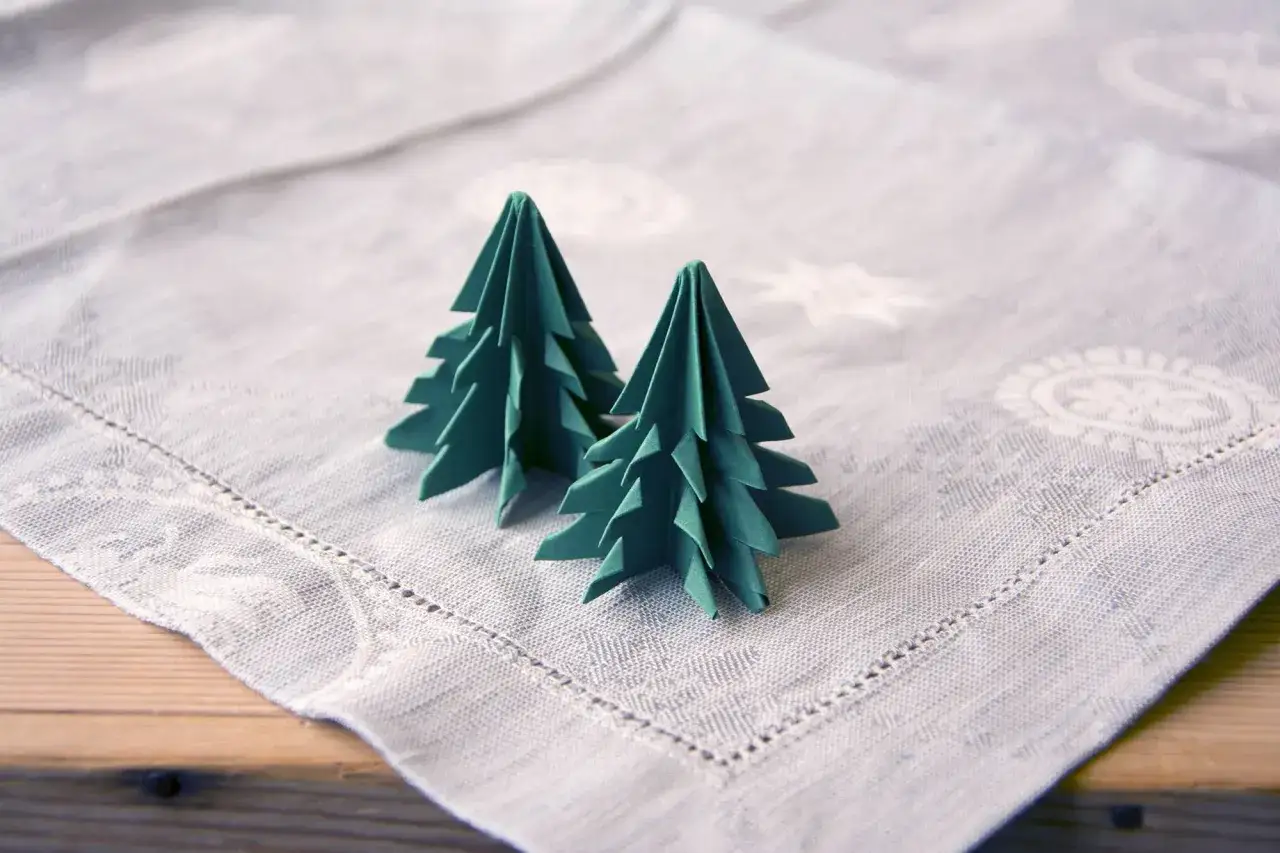 Zwei grüne Origami-Tannenbäume, die auf einer Tischdecke stehen. Eine einfache tannenbaum falten anleitung für Weihnachtsdekoration.