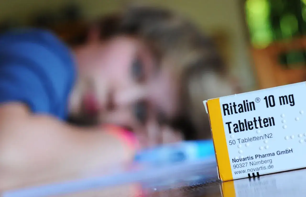 Nahaufnahme einer Packung Ritalin 10 mg Tabletten, im Hintergrund eine unscharfe Person.