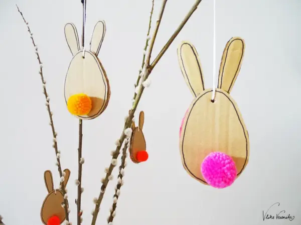 Osterhasen aus Pappe mit bunten Bommeln als Schwänzen, perfekt zum Basteln Ostern Kinder. Hängen an Weidenkätzchen-Zweigen.